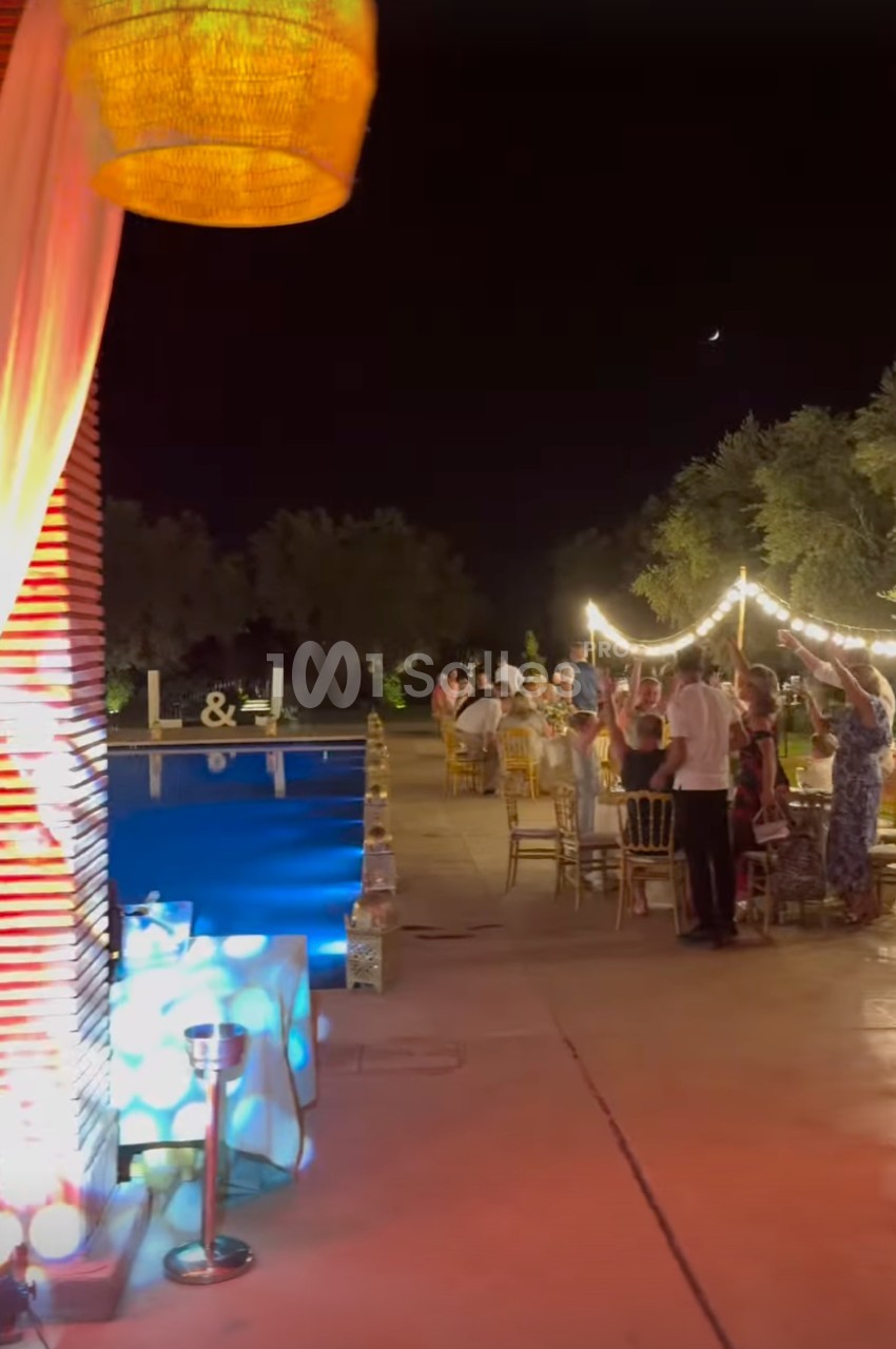 Des invités rassemblés autour de tables décorées sous des guirlandes lumineuses près d'une piscine, en soirée.