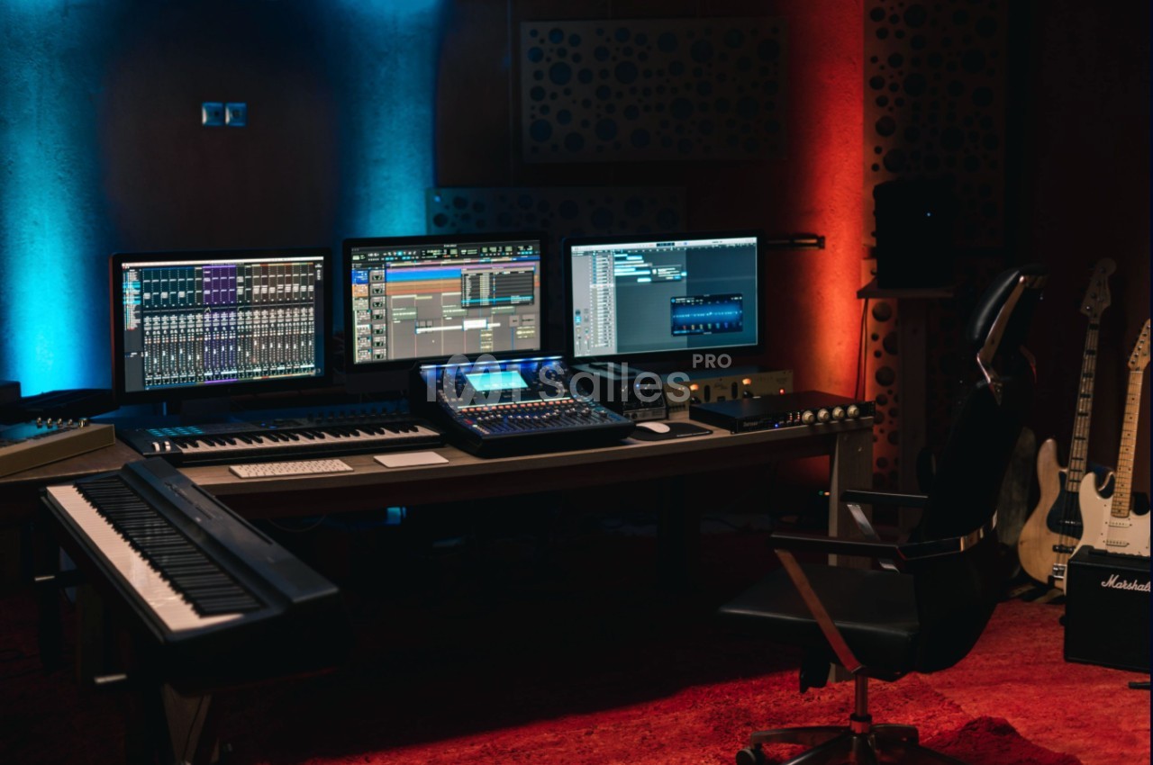 Poste de travail de studio d'enregistrement avec écrans, clavier, console audio et guitares dans une ambiance éclairée.