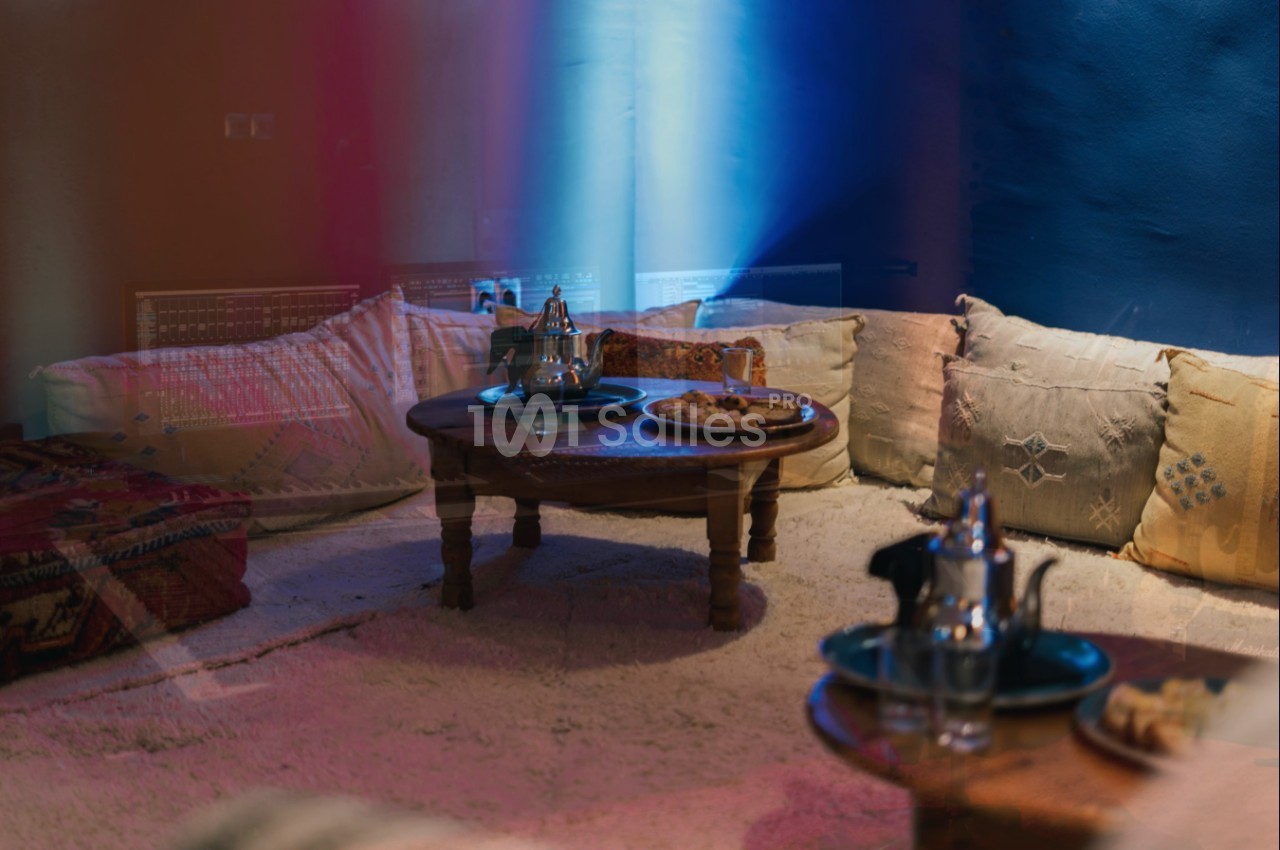 Salon marocain avec tables basses en bois, théières, plateaux et coussins colorés sur un tapis clair.