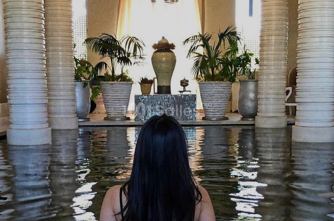 Une femme de dos dans un bassin intérieur entouré de colonnes, plantes et lumière naturelle.