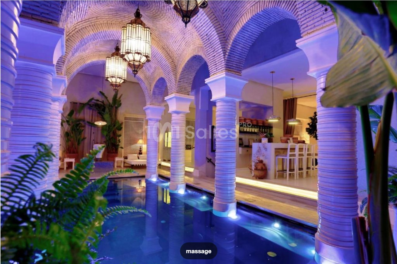 Intérieur d'un riad avec piscine éclairée, arches en pierre, luminaires suspendus et plantes tropicales.