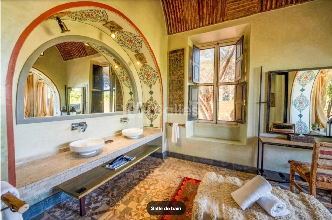 Salle de bain lumineuse avec double vasque, miroirs encadrés d'arcs décorés, fenêtre ouverte et tapis au sol.
