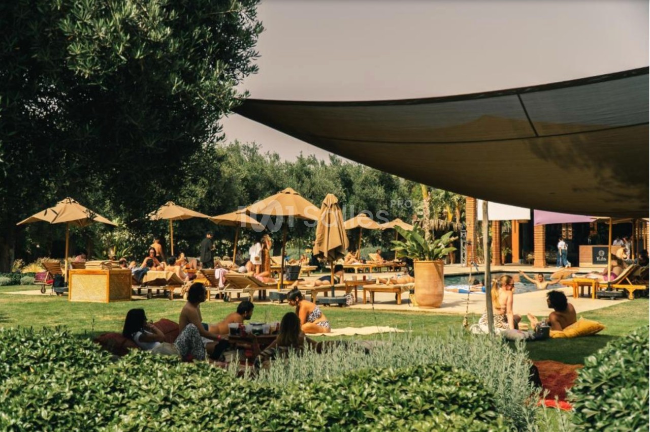 Personnes profitant d'un espace extérieur avec transats, parasols, végétation et une grande toile d'ombrage.