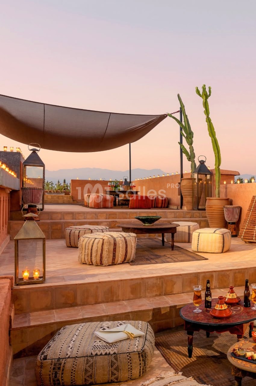 Terrasse aménagée avec coussins, tables basses, lanternes et vue sur un paysage au coucher du soleil.