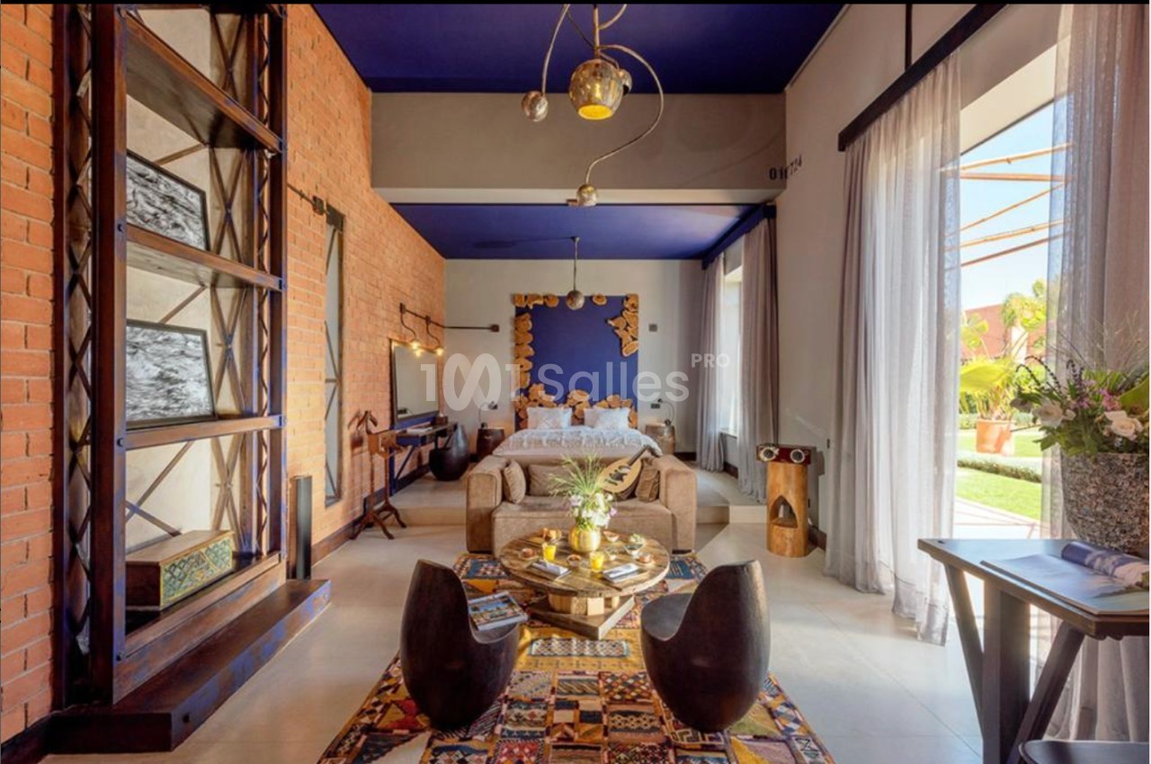 Chambre lumineuse avec murs en briques, plafond bleu, lit double, coin salon et grandes fenêtres donnant sur un jardin.