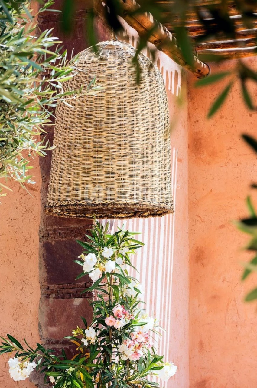 Suspension en osier tressé accrochée à un mur en stuc rose, entourée de plantes et de fleurs.