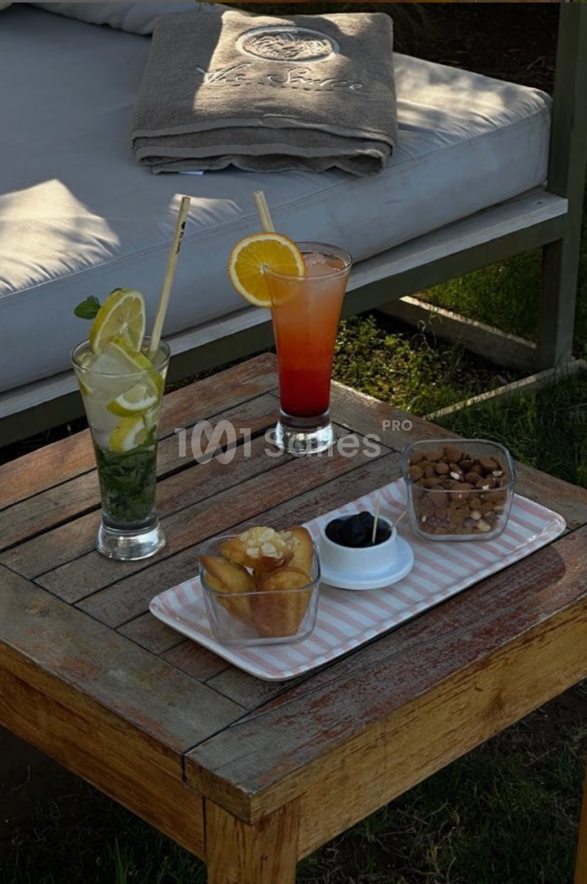 Boissons fraîches et collations variées sur une table en bois, près d'une banquette extérieure avec serviette pliée.
