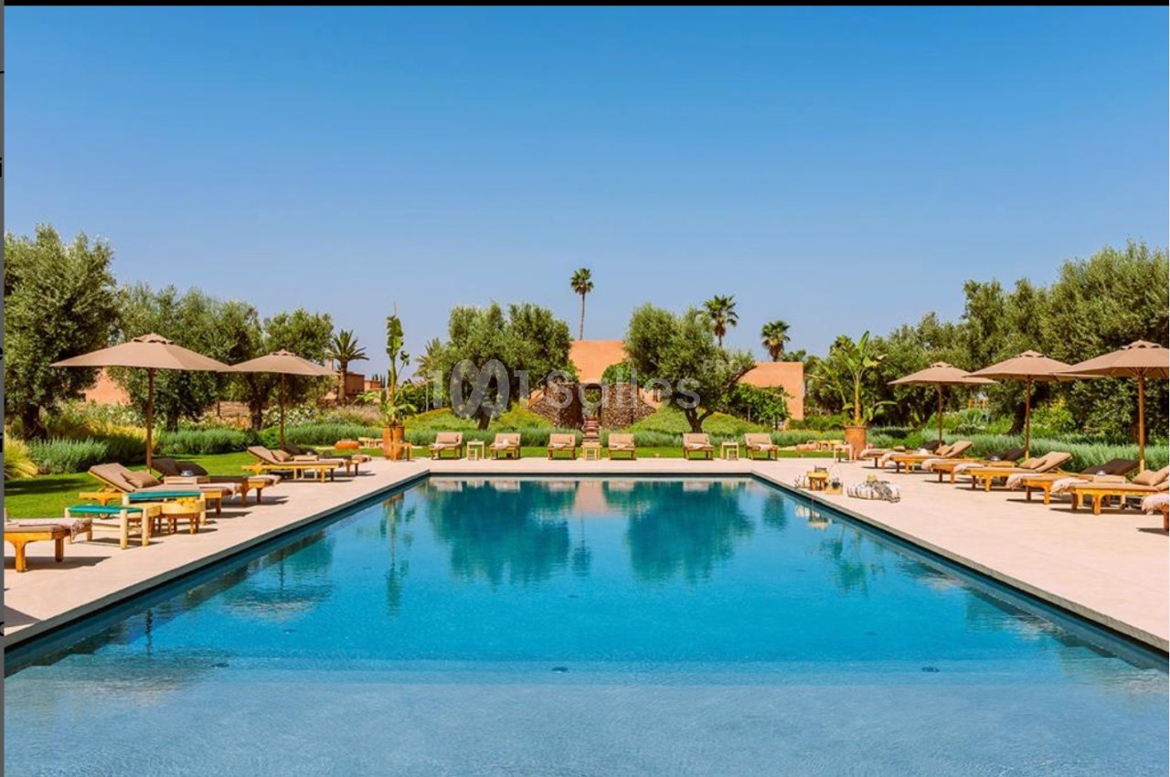 Piscine extérieure entourée de transats, parasols et végétation luxuriante sous un ciel bleu clair.
