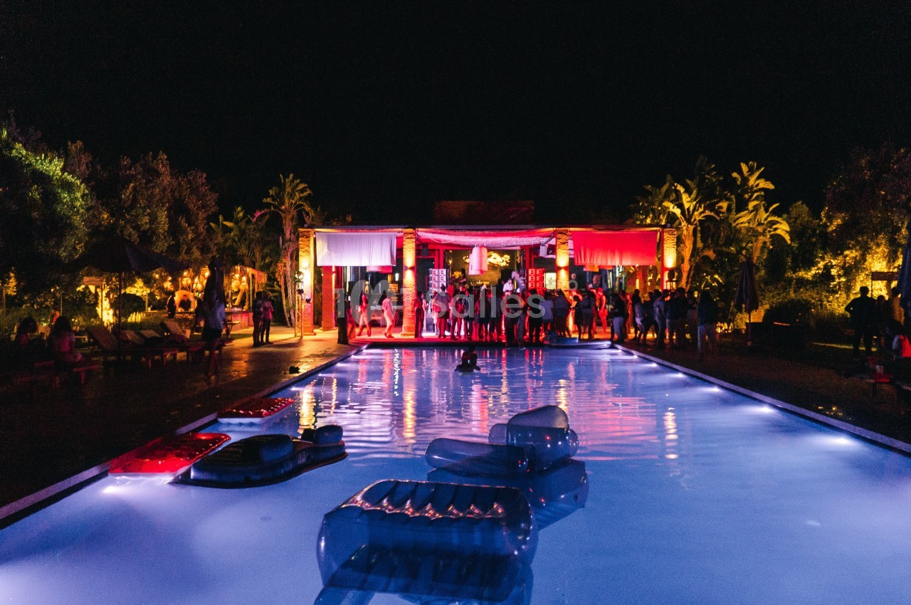 Soirée festive en extérieur avec des lumières colorées, une piscine éclairée et des invités rassemblés au fond.
