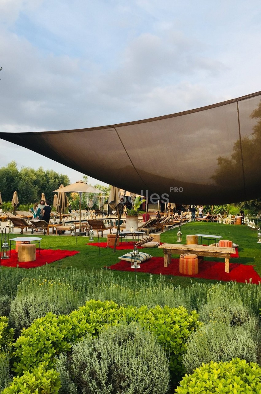 Espace extérieur aménagé avec des tapis rouges, des sièges bas, des parasols et une grande voile d'ombrage.