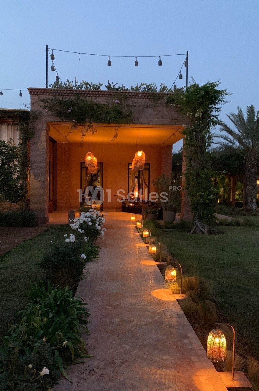 Allée éclairée par des lanternes menant à une maison en briques avec terrasse illuminée au crépuscule.