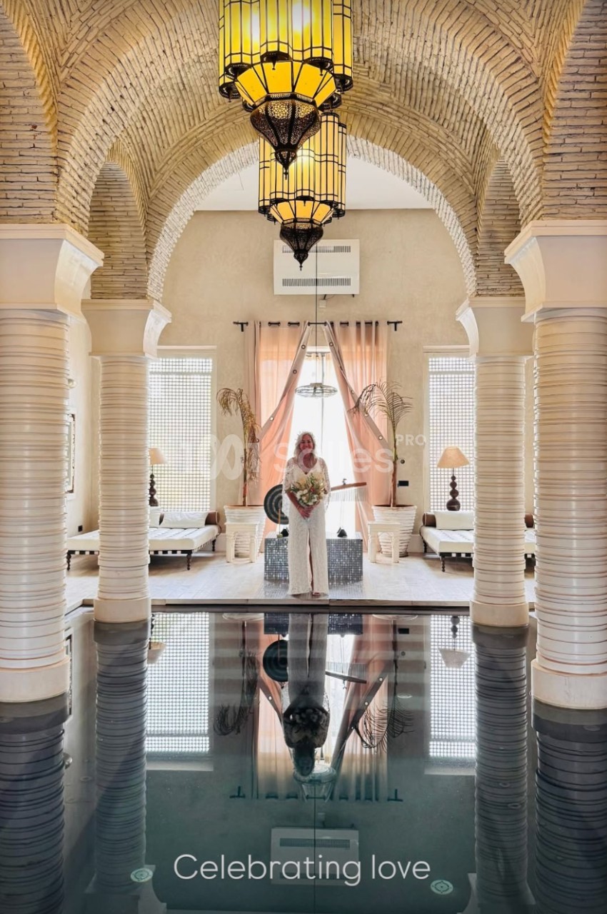 Une femme en robe blanche tient un bouquet dans une salle voûtée avec piscine intérieure et lumière naturelle.