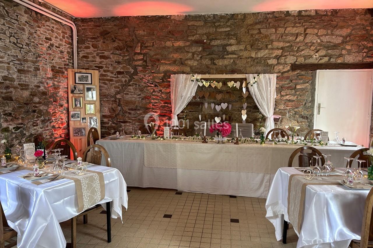 Salle décorée pour un événement, avec tables dressées, nappes blanches et un mur en pierre orné de cœurs suspendus.