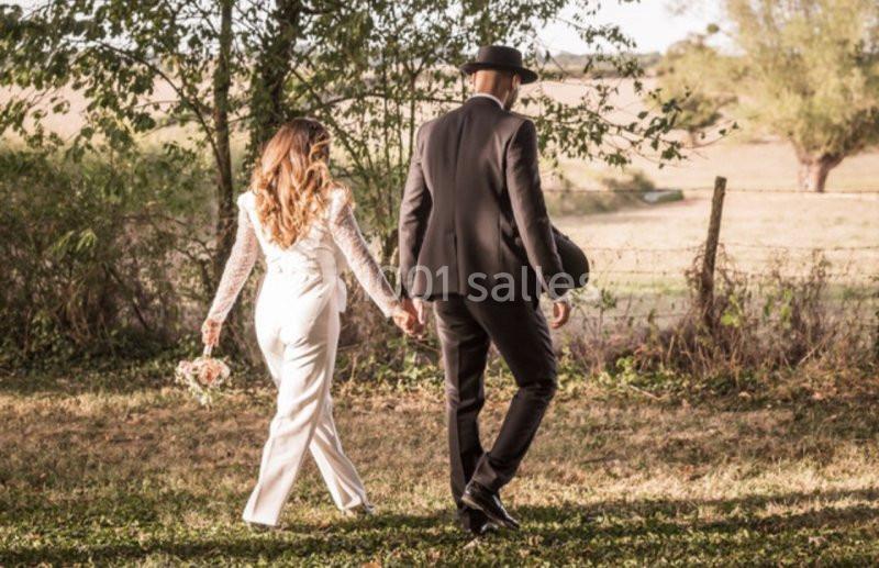 Location salle Maligny (Côte-d'Or) - Domaine du Grand Nanteux #21 Un couple élégant marche main dans la main dans un champ ensoleillé, entouré d'arbres et de verdure.