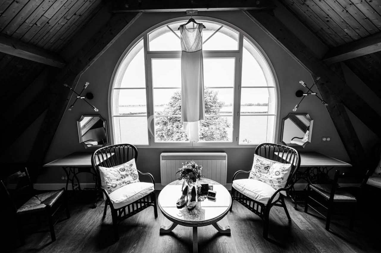 Location salle Maligny (Côte-d'Or) - Domaine du Grand Nanteux #33 Robe de mariée suspendue devant une grande fenêtre cintrée dans une pièce aménagée avec des fauteuils et une table basse.