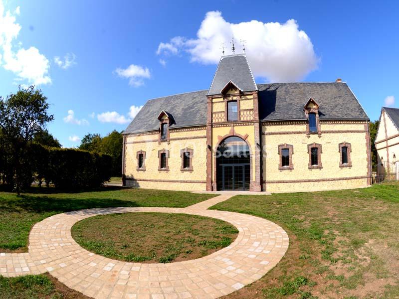 Location salle Sylvains-les-Moulins (Eure) - Domaine de Coulonge #5 Bâtiment en briques et pierres avec toit en ardoise, entouré d'une allée pavée en forme de cercle, sous un ciel bleu.