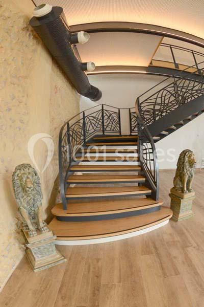 Location salle Sylvains-les-Moulins (Eure) - Domaine de Coulonge #19 Escalier en bois avec garde-corps en métal, encadré par deux statues de lions sur des socles, dans un intérieur moderne.