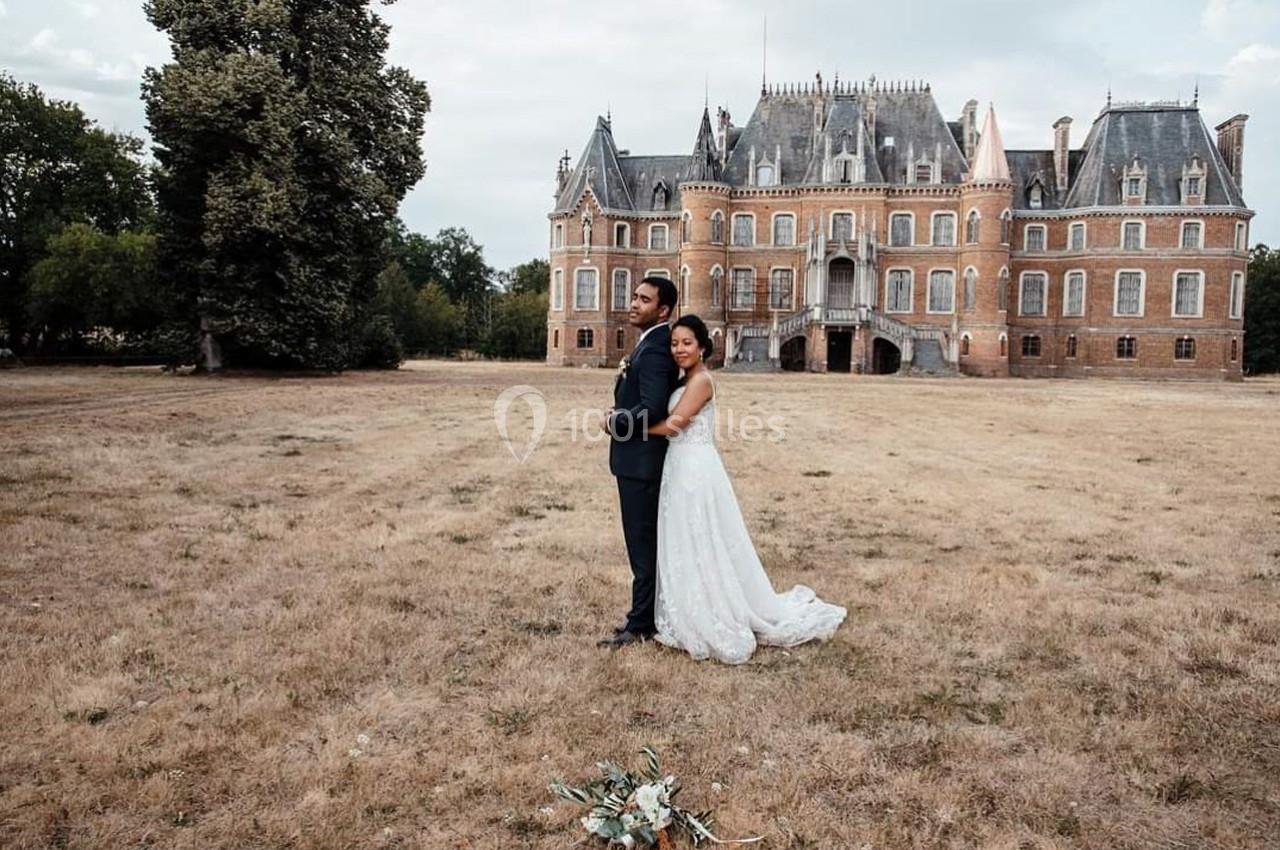 Location salle Sylvains-les-Moulins (Eure) - Domaine de Coulonge #28 Un couple en tenue de mariage pose devant un château entouré d'une pelouse et d'arbres.