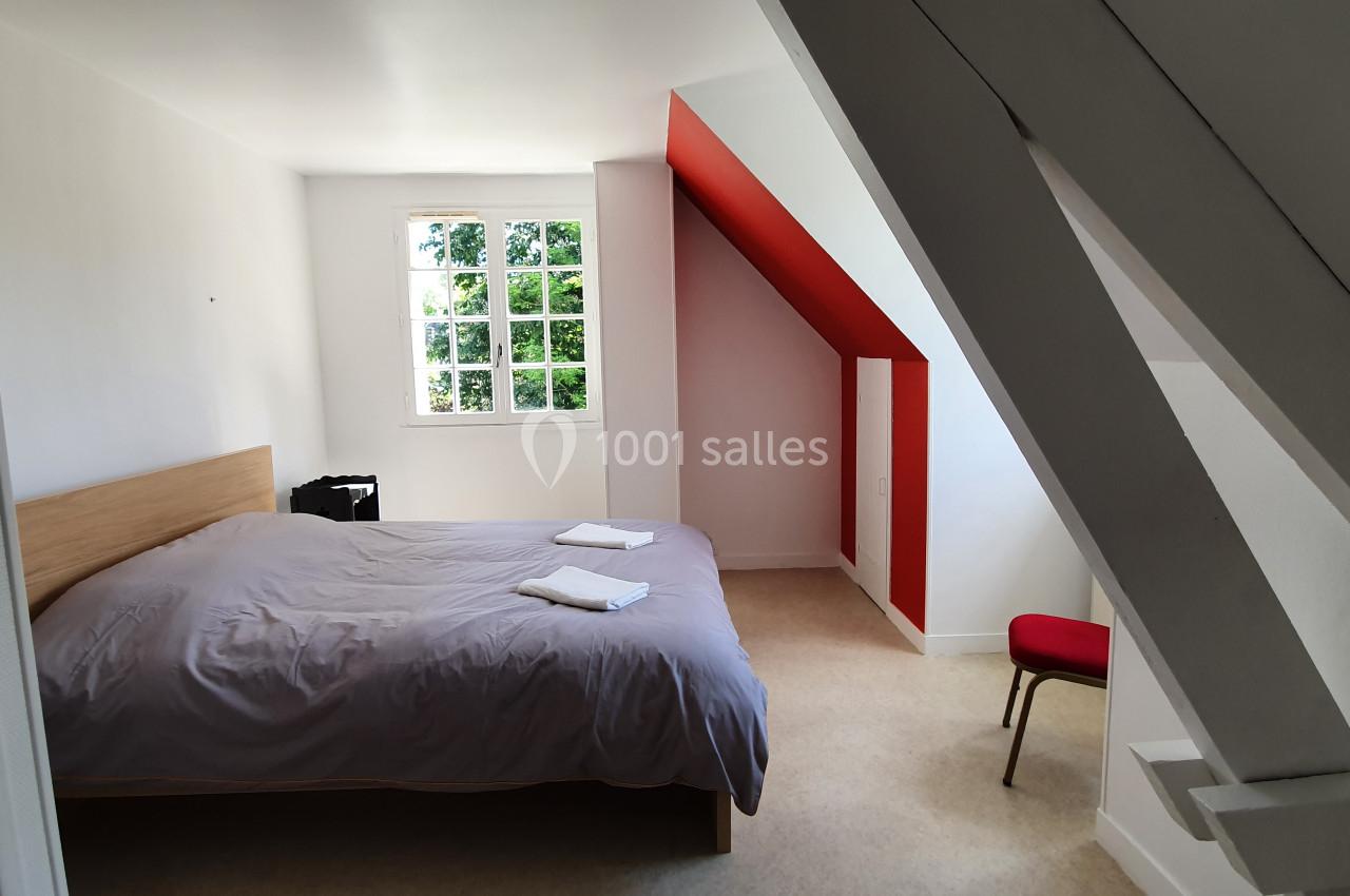 Location salle Sylvains-les-Moulins (Eure) - Domaine de Coulonge #36 Chambre mansardée lumineuse avec un lit double, draps gris, chaise rouge et fenêtre donnant sur un espace verdoyant.