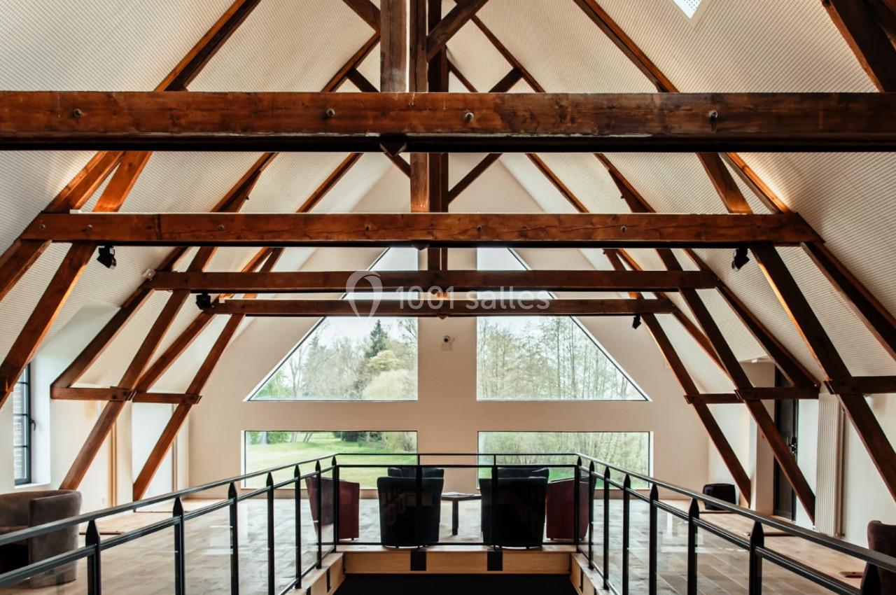 Location salle Sylvains-les-Moulins (Eure) - Domaine de Coulonge #25 Intérieur d'un bâtiment avec une charpente en bois apparente, mezzanine et grandes fenêtres laissant entrer la lumière…