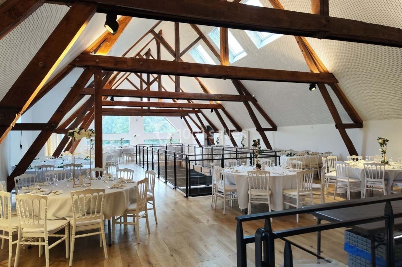 Location salle Sylvains-les-Moulins (Eure) - Domaine de Coulonge #20 Salle lumineuse avec poutres apparentes, tables rondes dressées pour un événement, parquet en bois clair et baies vitrées.