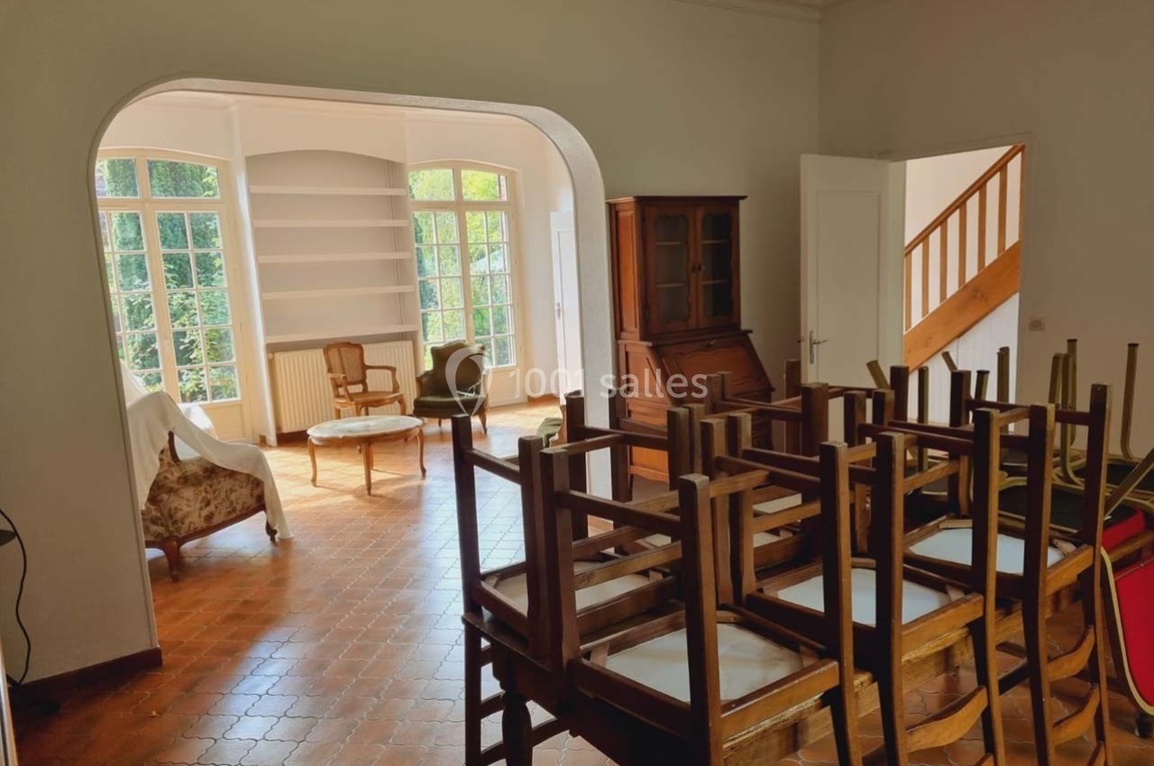 Location salle Sylvains-les-Moulins (Eure) - Domaine de Coulonge #38 Salle lumineuse avec chaises empilées, sol en terre cuite, meubles en bois et grandes fenêtres donnant sur un jardin.