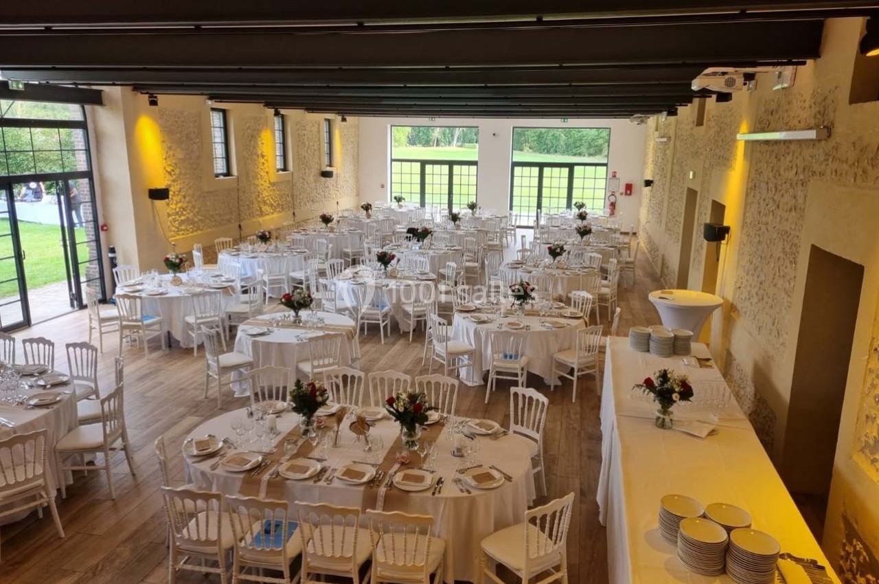 Location salle Sylvains-les-Moulins (Eure) - Domaine de Coulonge #9 Salle de réception lumineuse avec tables rondes dressées, chaises blanches et décorations florales, vue sur un jardin.