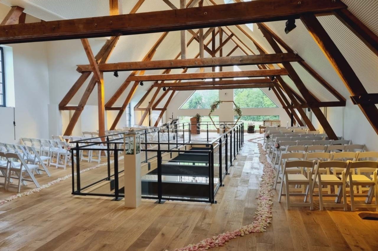 Location salle Sylvains-les-Moulins (Eure) - Domaine de Coulonge #23 Salle lumineuse avec poutres apparentes, chaises blanches alignées et allée décorée de pétales de fleurs.