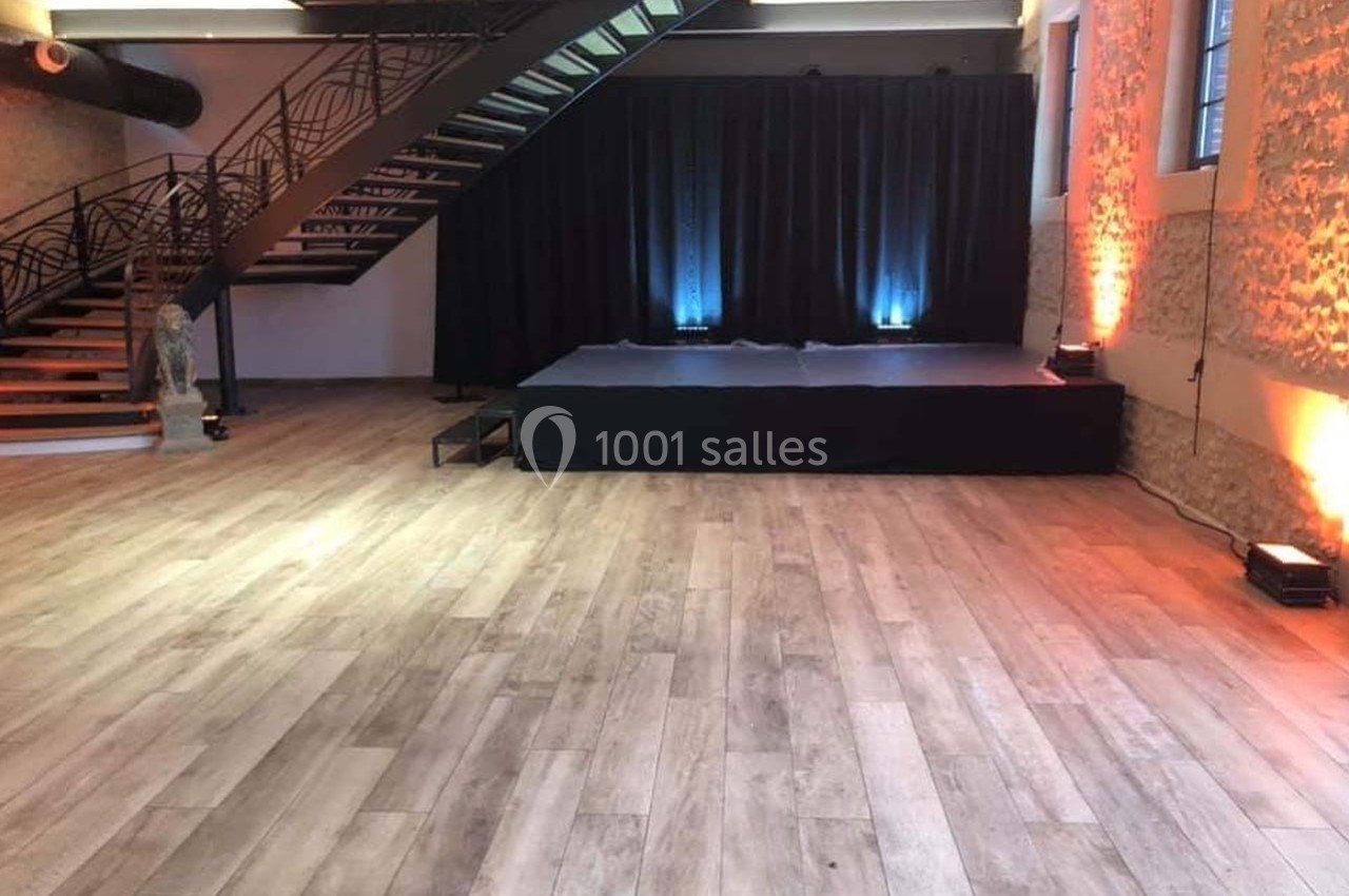 Location salle Sylvains-les-Moulins (Eure) - Domaine de Coulonge #52 Salle avec parquet clair, petite scène noire, escalier métallique et murs en pierre éclairés par des lumières colorées.