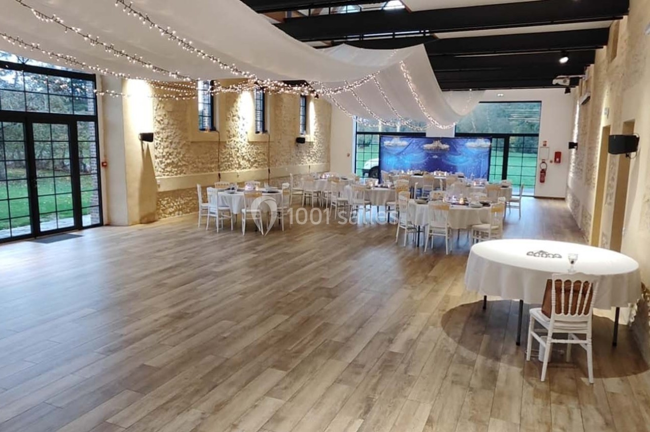 Location salle Sylvains-les-Moulins (Eure) - Domaine de Coulonge #4 Salle de réception lumineuse avec parquet, tables dressées, murs en pierre et grandes baies vitrées donnant sur un jardin.