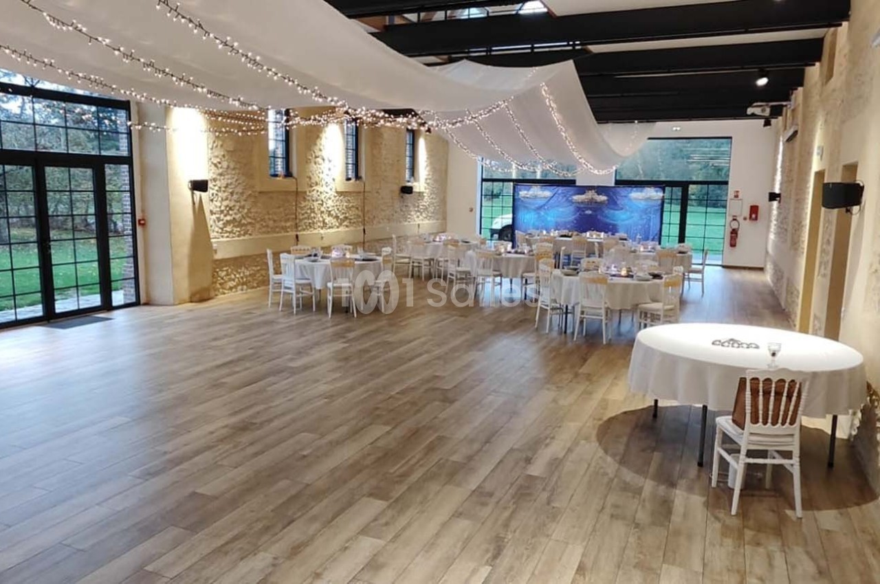 Salle de réception lumineuse avec parquet, tables dressées, murs en pierre et grandes baies vitrées donnant sur un jardin.