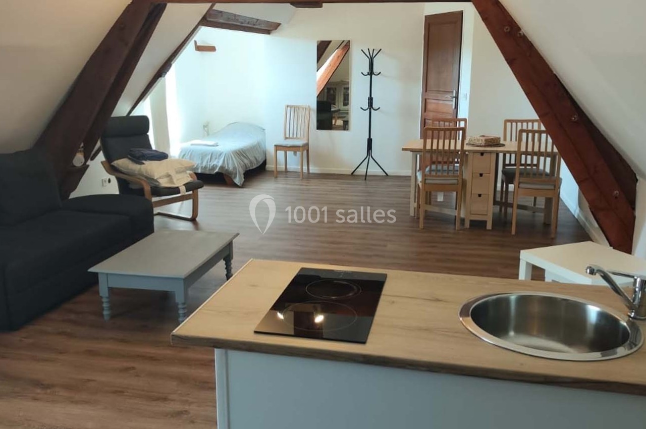 Location salle Sylvains-les-Moulins (Eure) - Domaine de Coulonge #40 Vue d'un studio mansardé avec coin cuisine, table à manger, canapé, lit simple et sol en parquet.