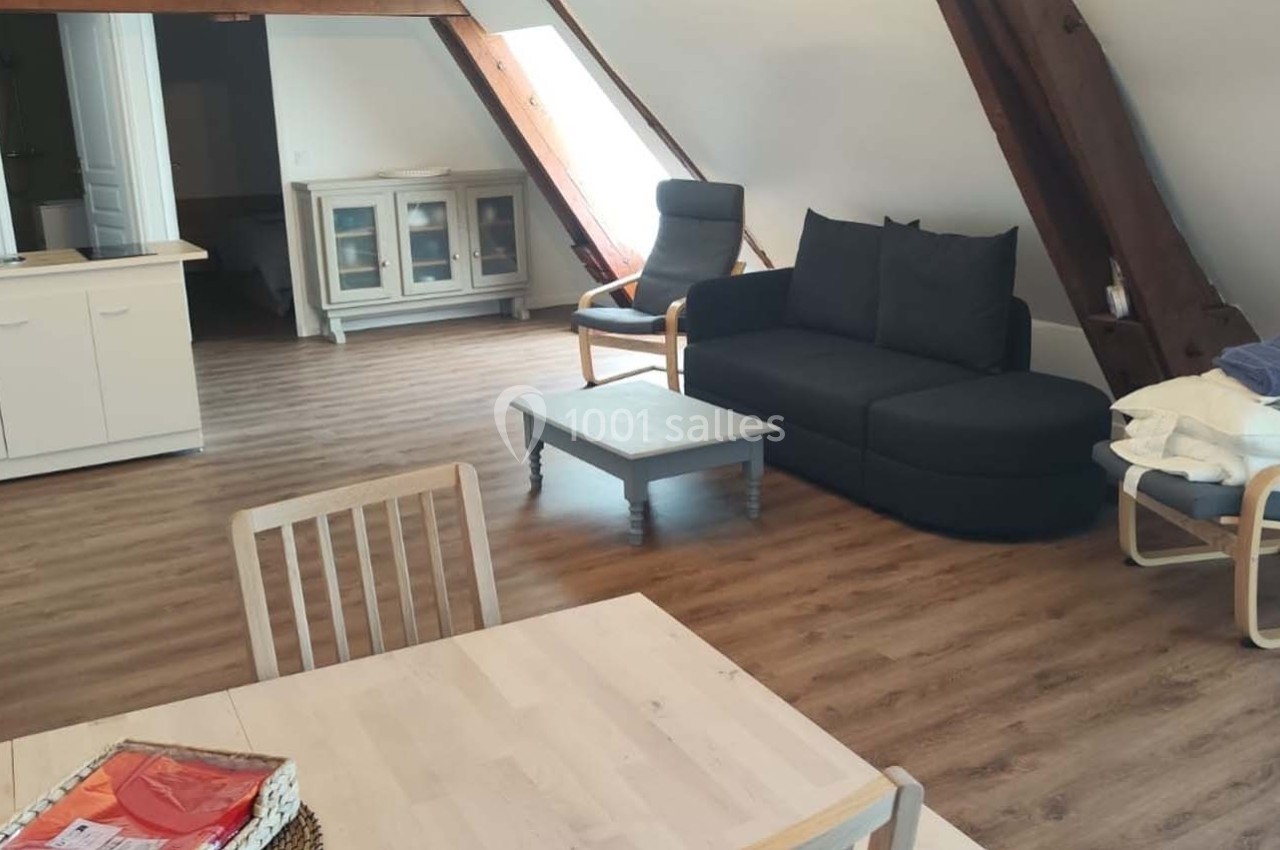 Location salle Sylvains-les-Moulins (Eure) - Domaine de Coulonge #41 Salon mansardé avec parquet, canapé noir, fauteuil, table basse, table à manger et cuisine ouverte en arrière-plan.