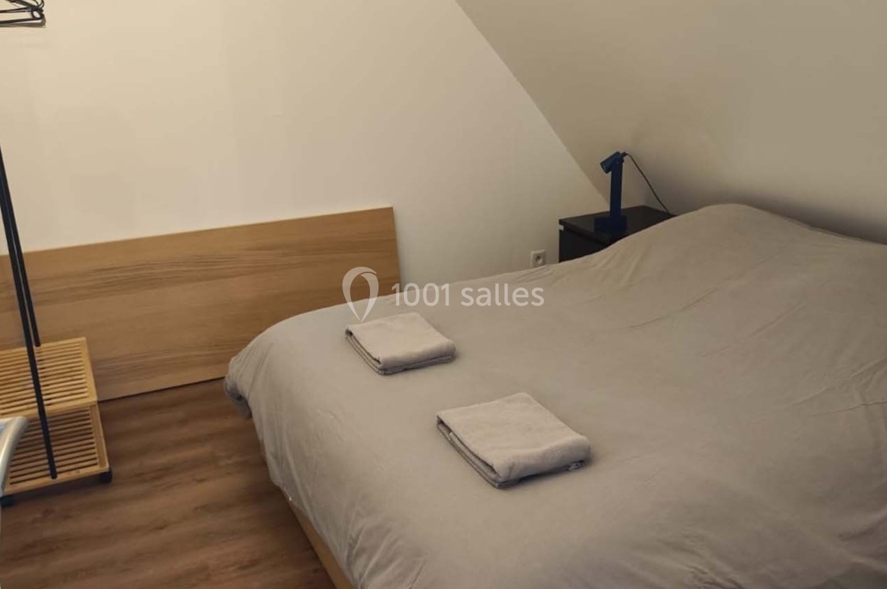 Location salle Sylvains-les-Moulins (Eure) - Domaine de Coulonge #45 Chambre mansardée avec lit double, draps gris, deux serviettes pliées, lampe de chevet et porte-manteau en métal.