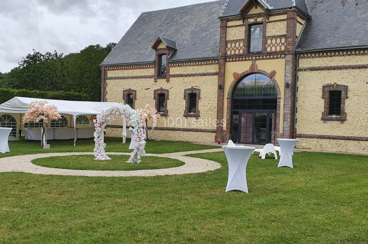 Location salle Sylvains-les-Moulins (Eure) - Domaine de Coulonge #50 Cour extérieure avec pelouse, arche fleurie, tables hautes et tente blanche devant un bâtiment en briques et pierre.