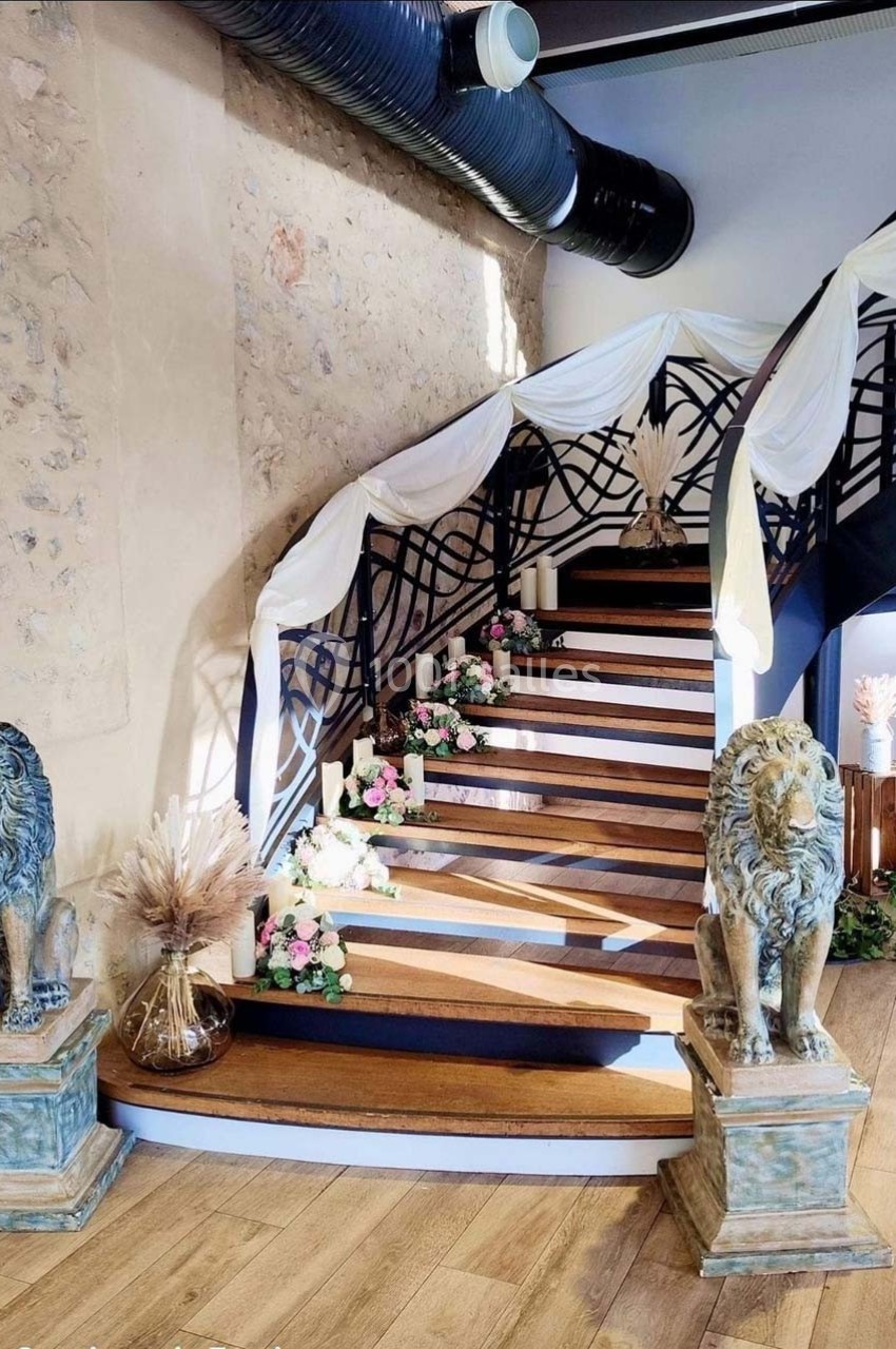 Location salle Sylvains-les-Moulins (Eure) - Domaine de Coulonge #17 Escalier en bois décoré de fleurs et drapés blancs, encadré par deux statues de lions dans une salle au mur en pierre.
