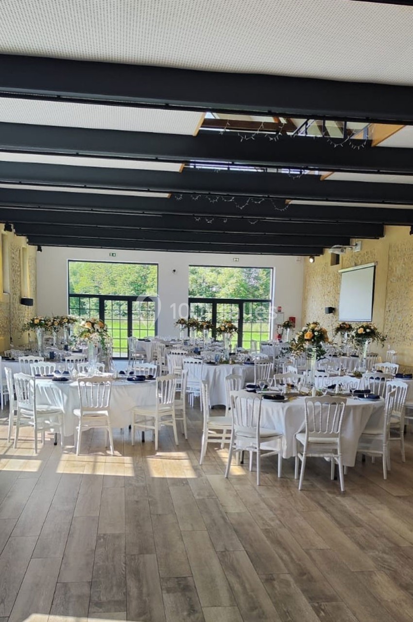 Location salle Sylvains-les-Moulins (Eure) - Domaine de Coulonge #15 Salle de réception lumineuse avec tables décorées, chaises blanches et grandes baies vitrées donnant sur un espace vert.
