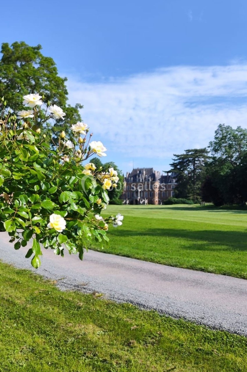 Location salle Sylvains-les-Moulins (Eure) - Domaine de Coulonge #2 Allée bordée de pelouse avec des roses blanches au premier plan et un château entouré d'arbres en arrière-plan.