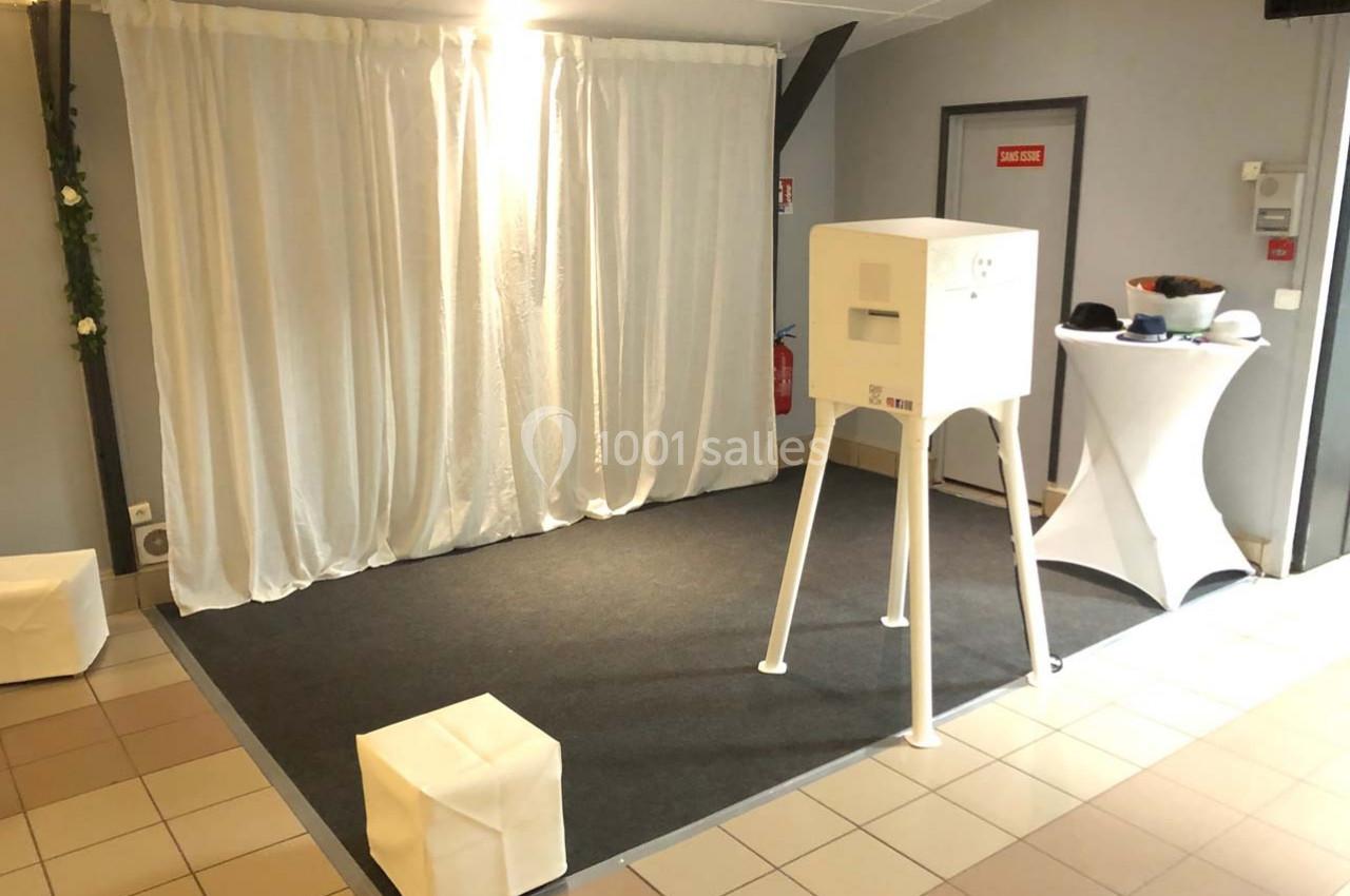 Espace photo avec cabine blanche sur trépied, rideau blanc en fond, table haute et tabourets dans une salle éclairée.