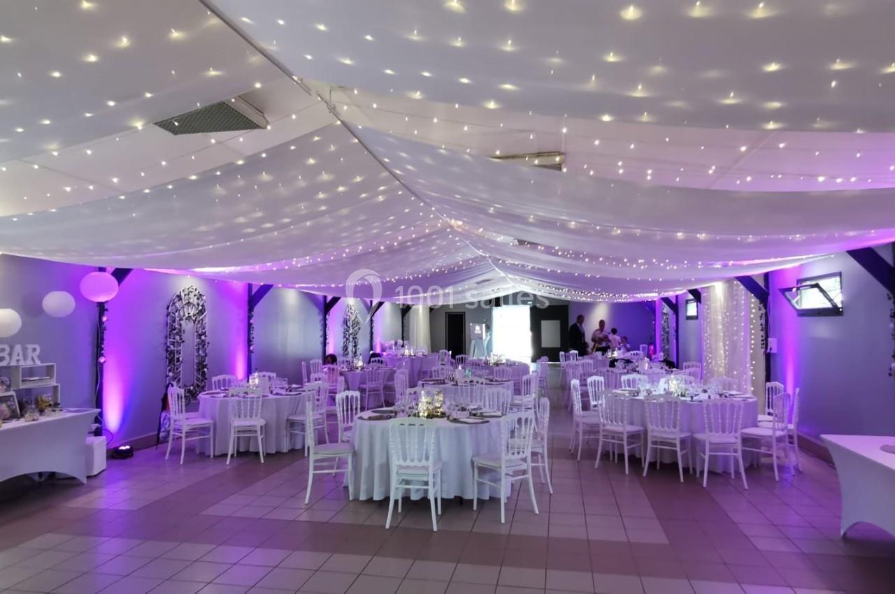 Salle de réception décorée avec des guirlandes lumineuses au plafond, tables rondes dressées et éclairage violet.