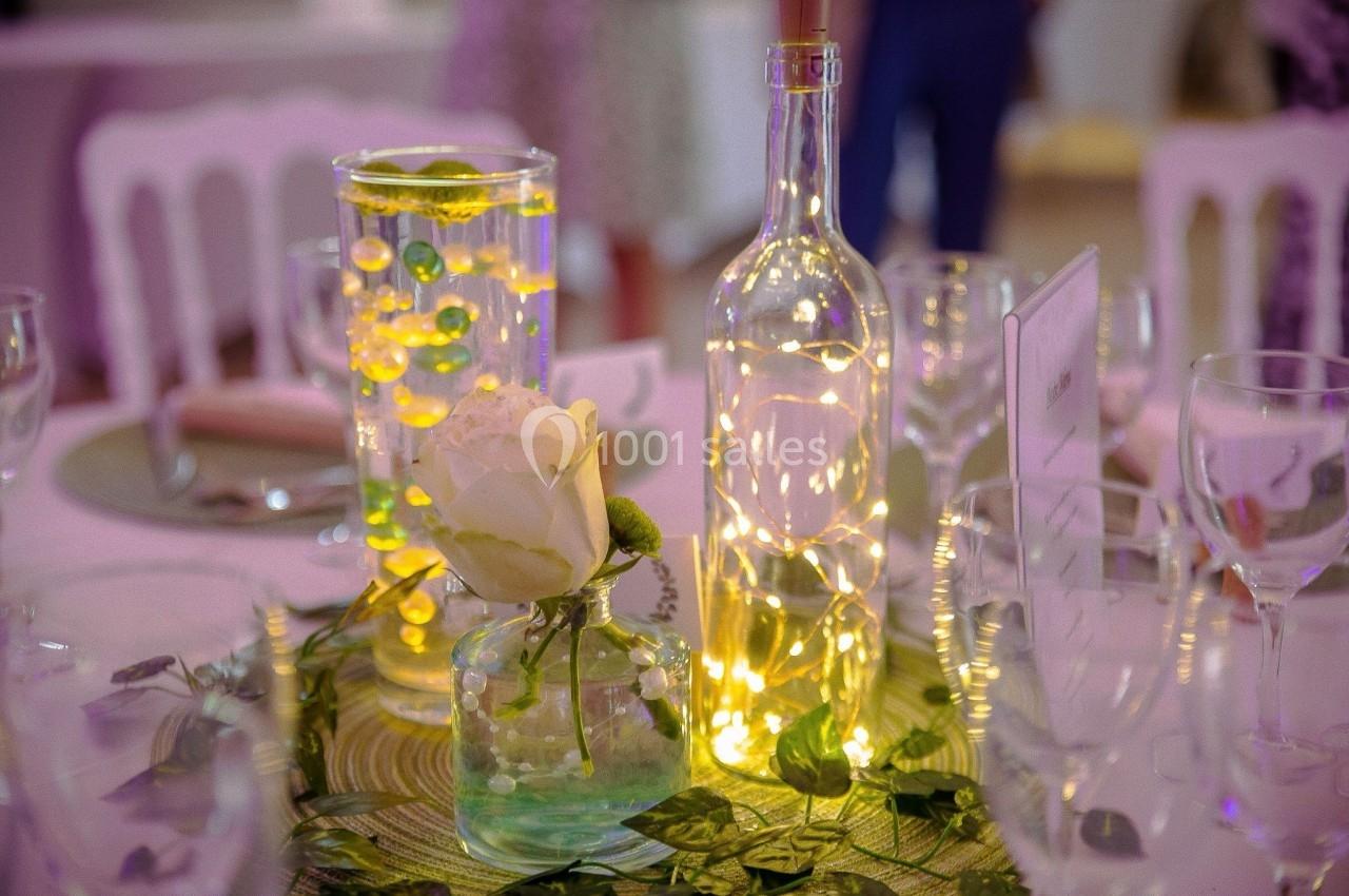 Centre de table décoratif avec une rose blanche, des guirlandes lumineuses et des vases en verre sur une nappe verte.
