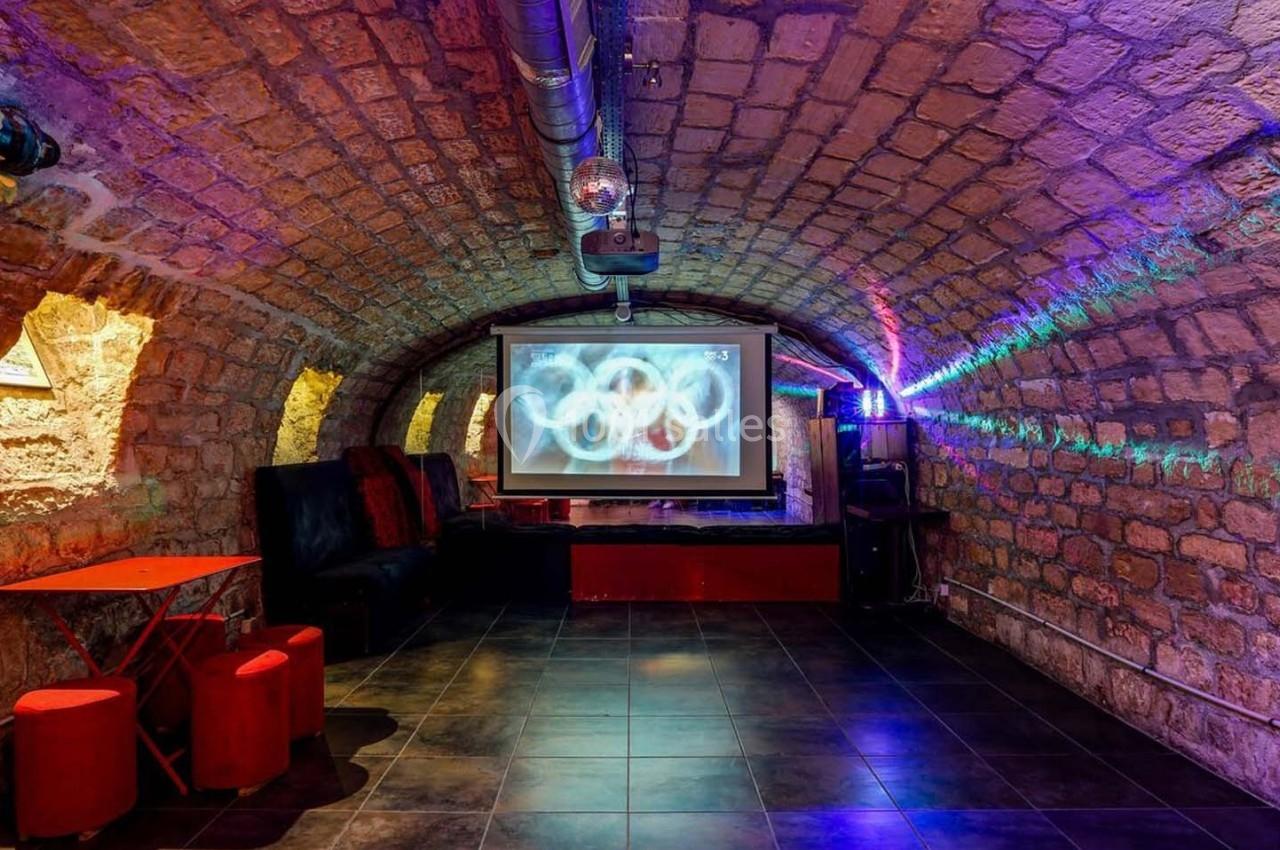 Salle voûtée en pierre avec un écran de projection, mobilier rouge et éclairage coloré.
