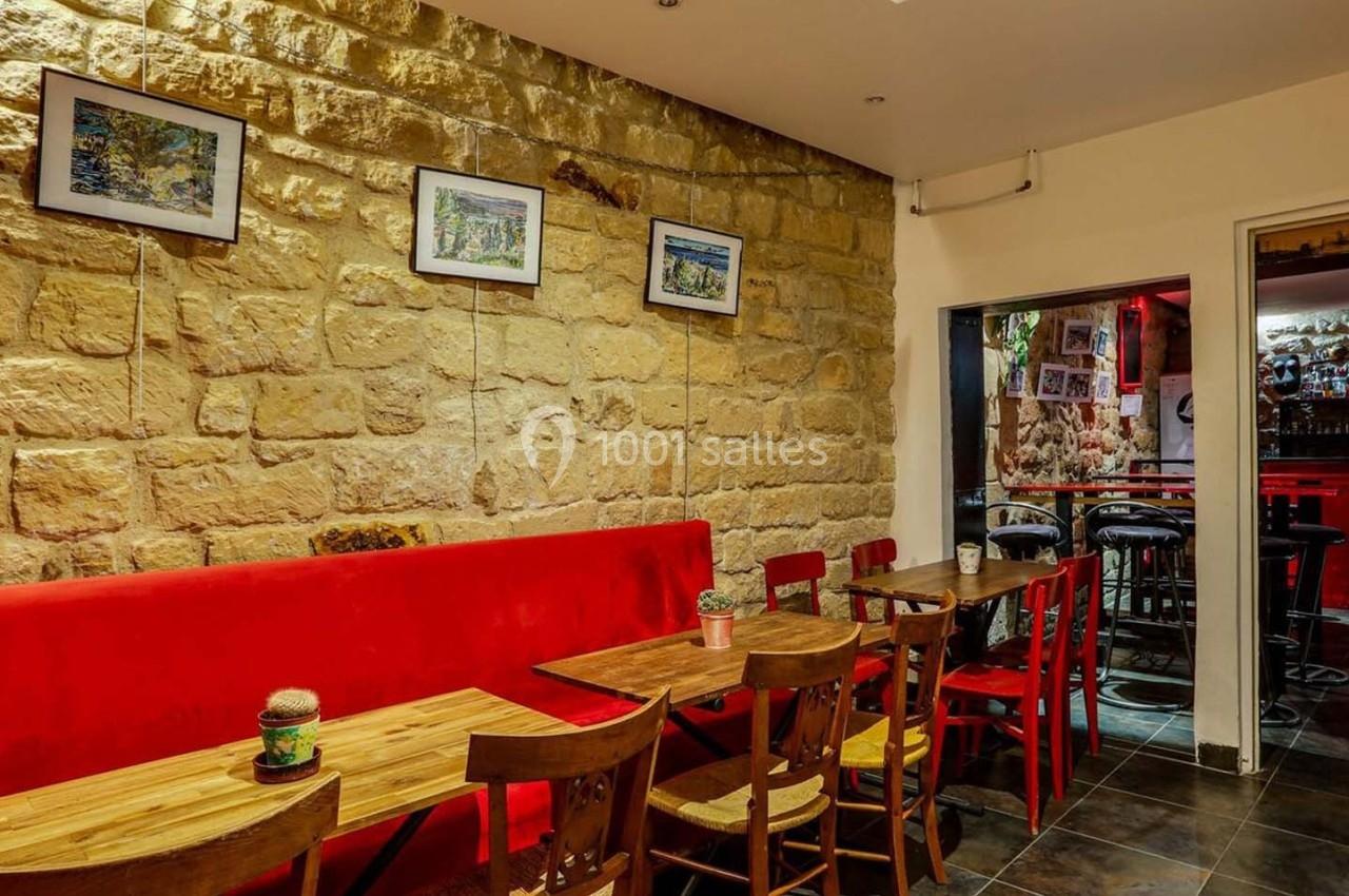 Salle de restaurant avec murs en pierre, banquette rouge, tables en bois et vue sur un bar éclairé en rouge.