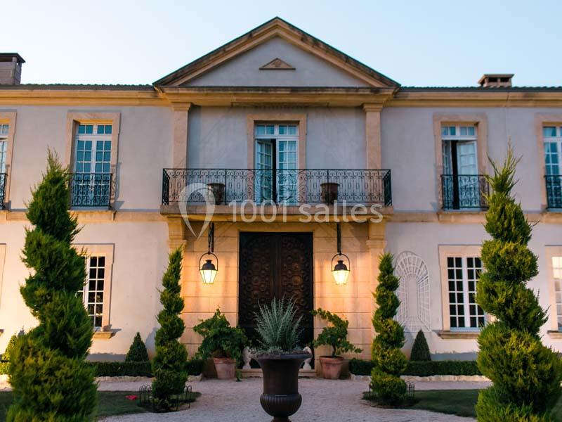 Façade d'une grande maison élégante avec balcon, fenêtres symétriques et allée bordée d'arbustes taillés.