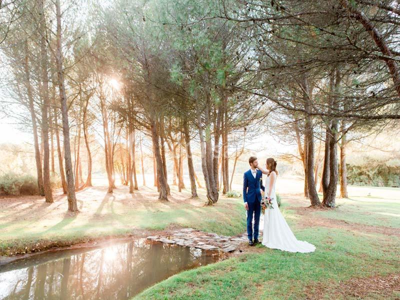 Un couple en tenue de mariage se tient près d'un petit pont dans une clairière boisée baignée de lumière.