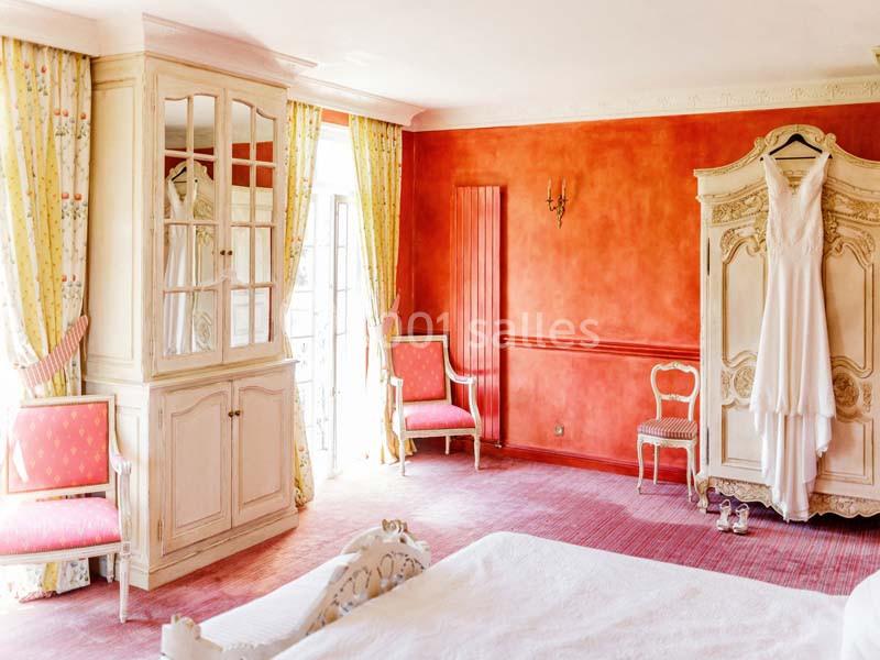 Chambre lumineuse avec murs orange, armoire blanche, robe de mariée suspendue et fauteuils roses près de fenêtres.