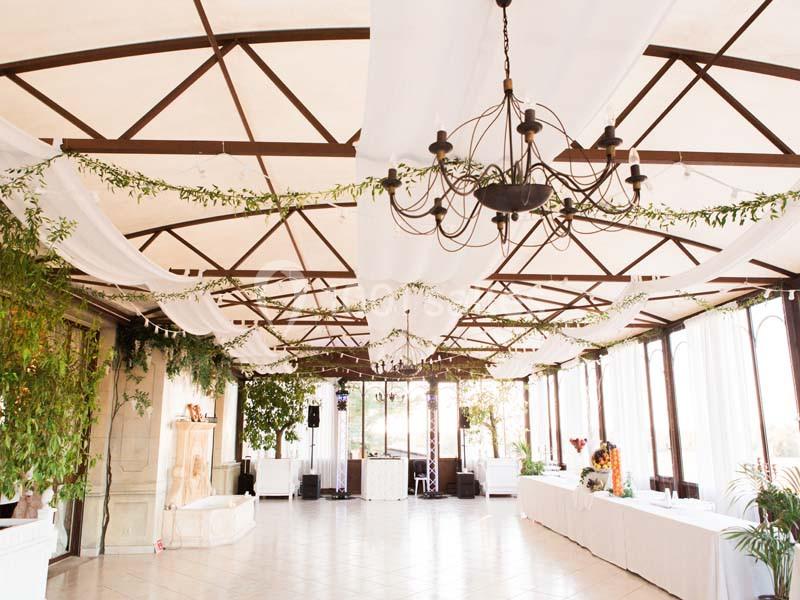 Salle lumineuse décorée de guirlandes végétales, de voilages blancs et d'un lustre suspendu au plafond.