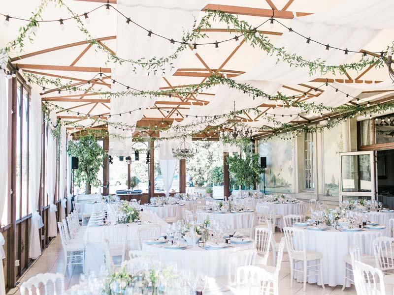 Salle lumineuse décorée pour un événement, avec tables rondes dressées, chaises blanches et guirlandes suspendues.
