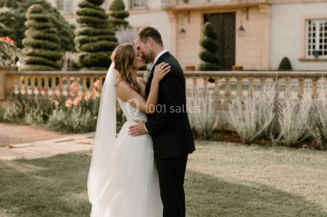 Un couple en tenue de mariage s'embrasse dans un jardin devant une grande maison élégante.