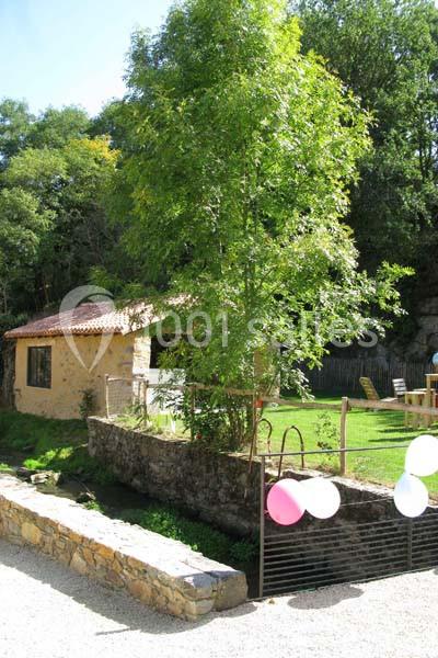 Petit jardin avec pelouse, arbre, maison en pierre, clôture et ballons accrochés à un portail métallique.